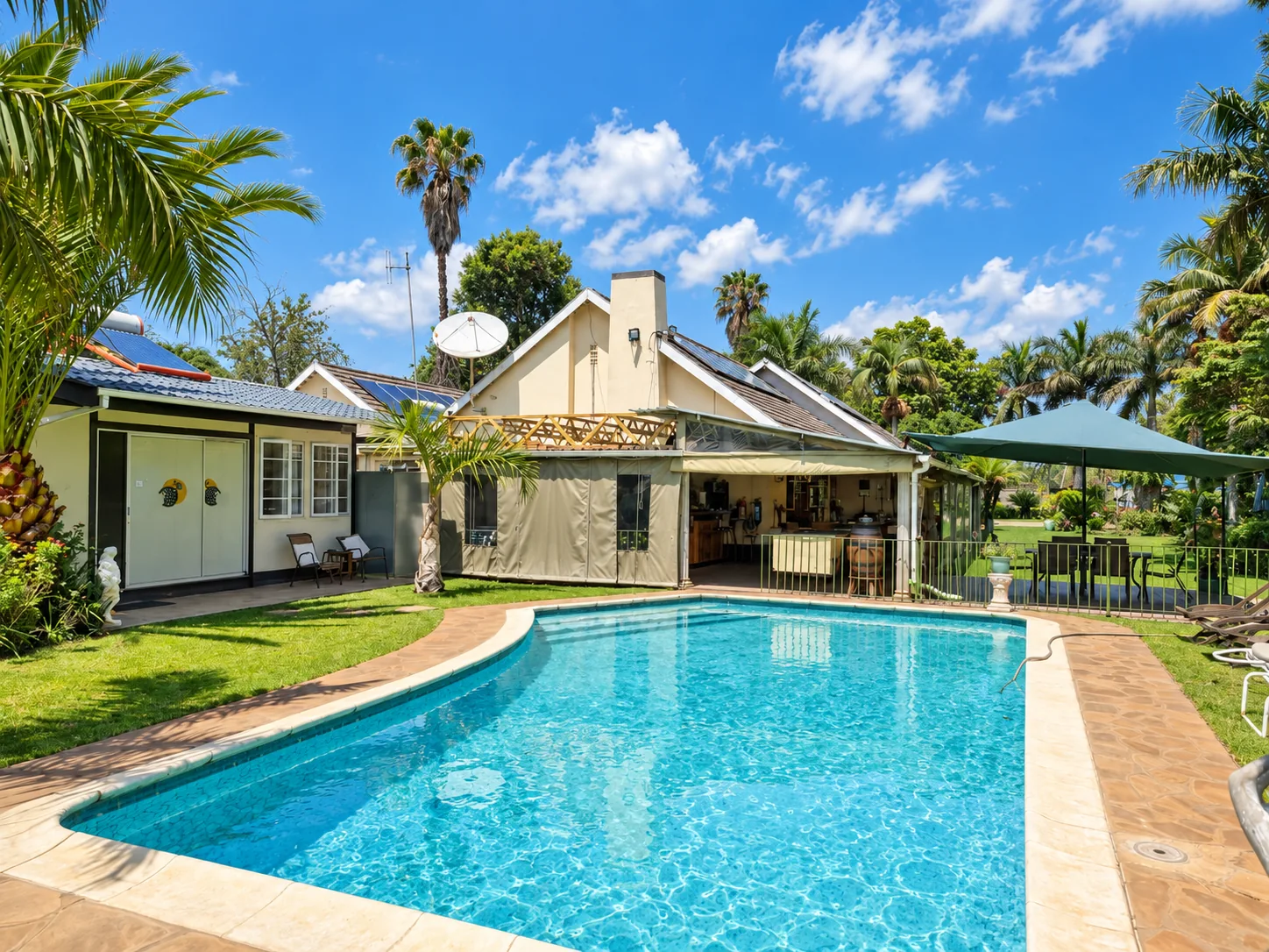 A blue outdoor swimming pool surrounded by palms, lawns and the guesthouse