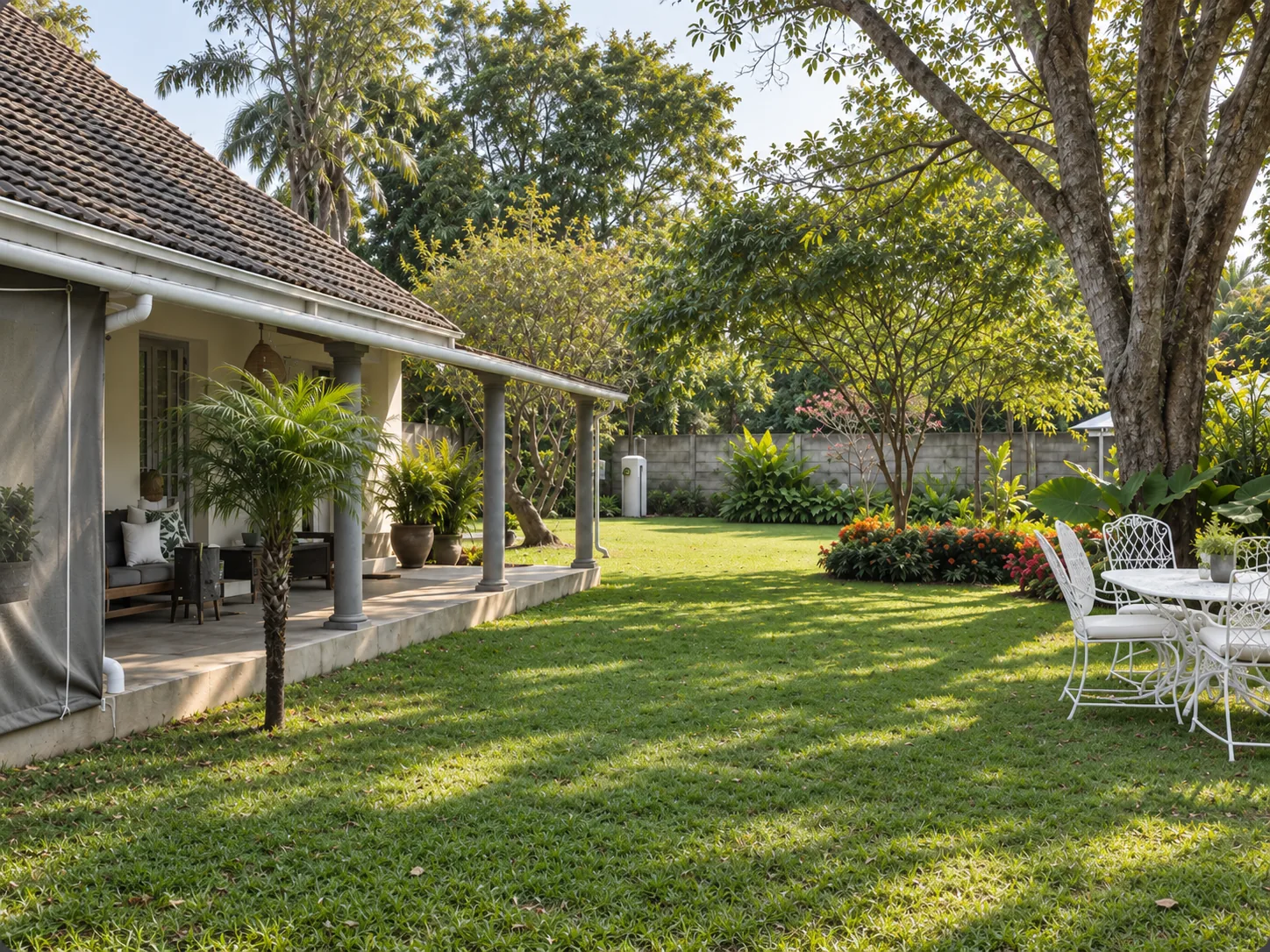 A tiled-roof guesthouse with a columned veranda looking onto a green lawn, garden furniture and mature trees